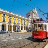 Czerwony tramwaj w Lizbonie, obok żółtego budynku i łuku triumfalnego. Czy do Portugalii potrzebny jest paszport? Tak, jeśli nie jesteś obywatelem UE.