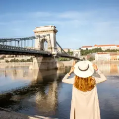 Kobieta w kapeluszu podziwia Most Łańcuchowy w Budapeszcie. W tle widok na Dunaj i budynki, a w portfelu czeka budapeszt waluta na dalsze zwiedzanie.
