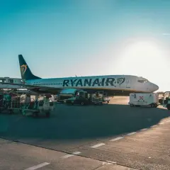Samolot Ryanair na płycie lotniska, przygotowywany do lotu. Widać wózki z bagażem i sprzęt obsługi naziemnej.