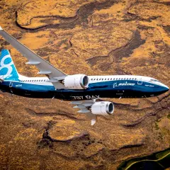 Boeing 737 MAX 8-200 z niebieskim ogonem z cyfrą 8 leci nad pustynnym krajobrazem.