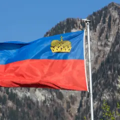 Flaga Liechtensteinu powiewa na tle gór. W jej centrum widnieje złota korona, symbol tego kraju i jego waluty.