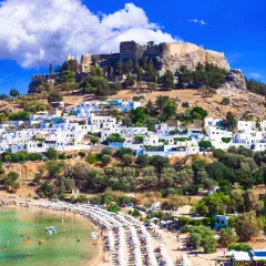 Widok na Lindos na Rodos z zamkiem na wzgórzu i plażą pełną ludzi. Woda jest krystalicznie czysta, idealna do kąpieli, co sugeruje przyjemną rodos temperaturę wody.