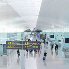 Jasny, przestronny terminal lotniska Barcelona El Prat. Pasażerowie poruszają się po nowoczesnym wnętrzu, kierując się znakami.