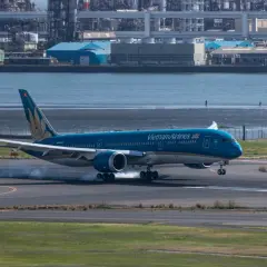 Błękitny 787 Dreamliner Vietnam Airlines ląduje, unosząc dym z opon na pasie startowym. W tle widać przemysłowy krajobraz i wodę.