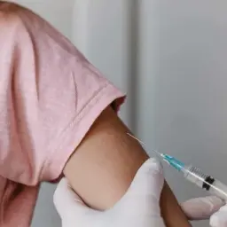 Dziecko w różowej koszulce otrzymuje szczepienie. Dłoń w rękawiczce trzyma strzykawkę. Tureckie szczepienia dla zdrowia.