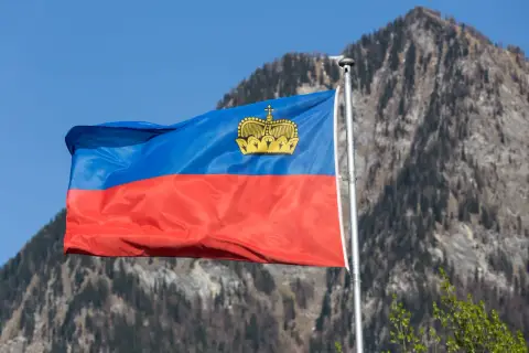 Flaga Liechtensteinu powiewa na tle gór. W jej centrum widnieje złota korona, symbol tego kraju i jego waluty.