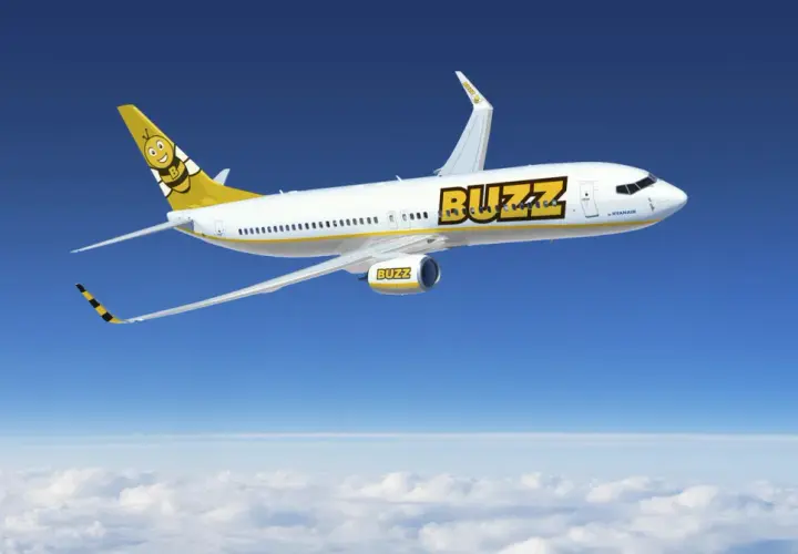Samolot Buzz, należący do Ryanair, leci nad chmurami. Słońce odbija się od białego kadłuba, a na ogonie widnieje uśmiechnięta pszczoła. Idealny na wakacje z bagażem!