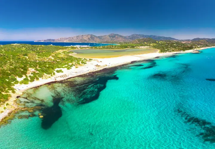 Widok z lotu ptaka na turkusowe morze i piaszczyste plaże w okolicach Cagliari. W oddali widać góry i zatokę z łodziami.