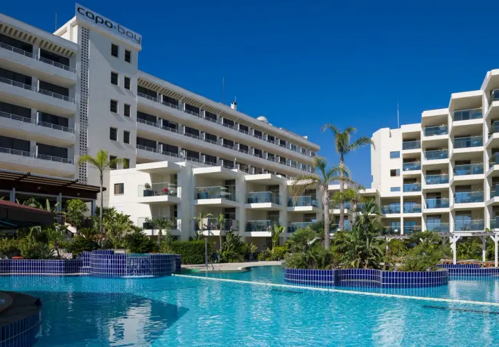 Basen hotelowy Capo Bay w Protaras z palmami i białymi budynkami hotelowymi pod błękitnym niebem.