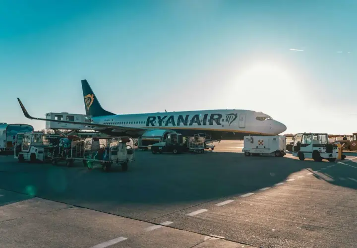 Samolot Ryanair na płycie lotniska, przygotowywany do lotu. Widać wózki z bagażem i sprzęt obsługi naziemnej.