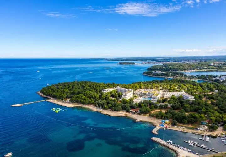 Hotel Delfin Plava Laguna z lotu ptaka, otoczony zielenią i błękitnym morzem. Widoczne baseny, plaża i przystań z łodziami.
