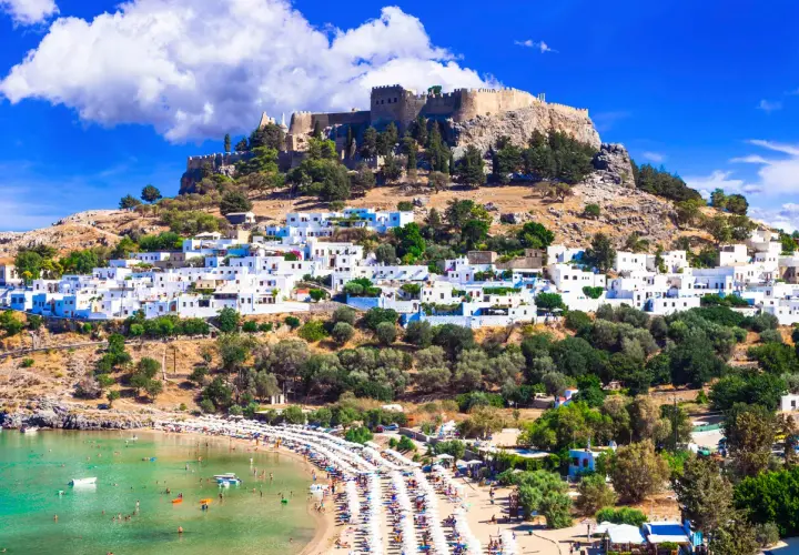 Widok na Lindos na Rodos z zamkiem na wzgórzu i plażą pełną ludzi. Woda jest krystalicznie czysta, idealna do kąpieli, co sugeruje przyjemną rodos temperaturę wody.