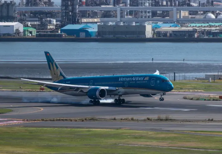 Błękitny 787 Dreamliner Vietnam Airlines ląduje, unosząc dym z opon na pasie startowym. W tle widać przemysłowy krajobraz i wodę.
