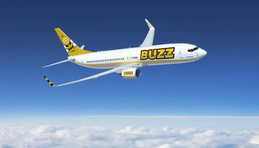 Samolot Buzz, należący do Ryanair, leci nad chmurami. Słońce odbija się od białego kadłuba, a na ogonie widnieje uśmiechnięta pszczoła. Idealny na wakacje z bagażem!