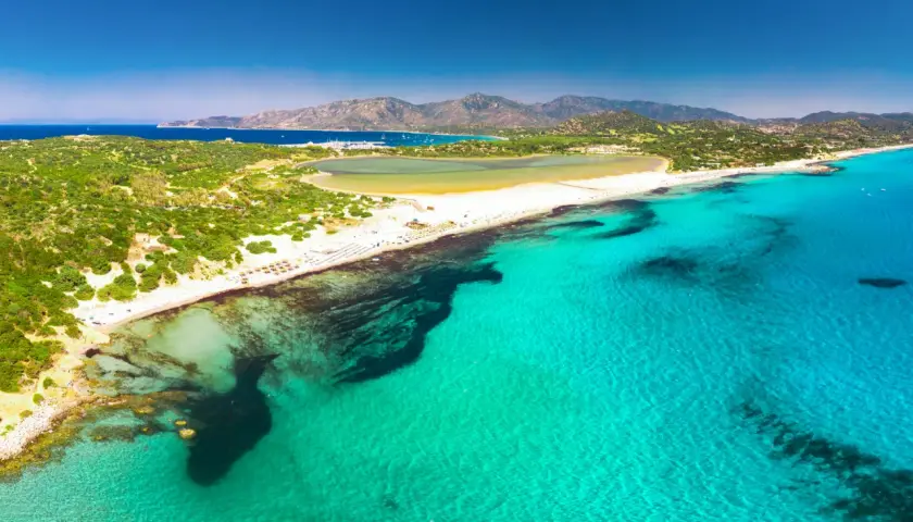 Widok z lotu ptaka na turkusowe morze i piaszczyste plaże w okolicach Cagliari. W oddali widać góry i zatokę z łodziami.