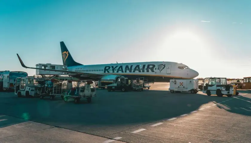 Samolot Ryanair na płycie lotniska, przygotowywany do lotu. Widać wózki z bagażem i sprzęt obsługi naziemnej.