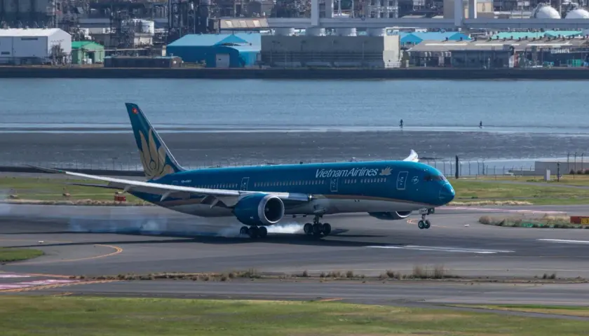 Błękitny 787 Dreamliner Vietnam Airlines ląduje, unosząc dym z opon na pasie startowym. W tle widać przemysłowy krajobraz i wodę.