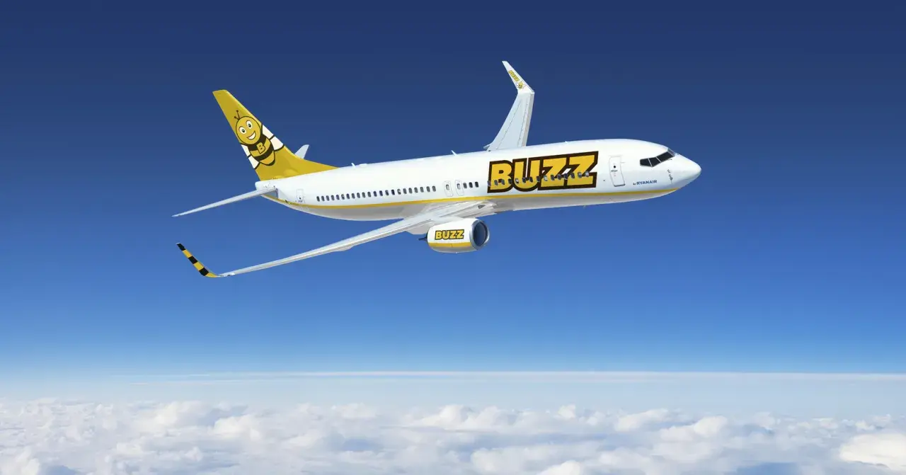 Samolot Buzz, należący do Ryanair, leci nad chmurami. Słońce odbija się od białego kadłuba, a na ogonie widnieje uśmiechnięta pszczoła. Idealny na wakacje z bagażem!
