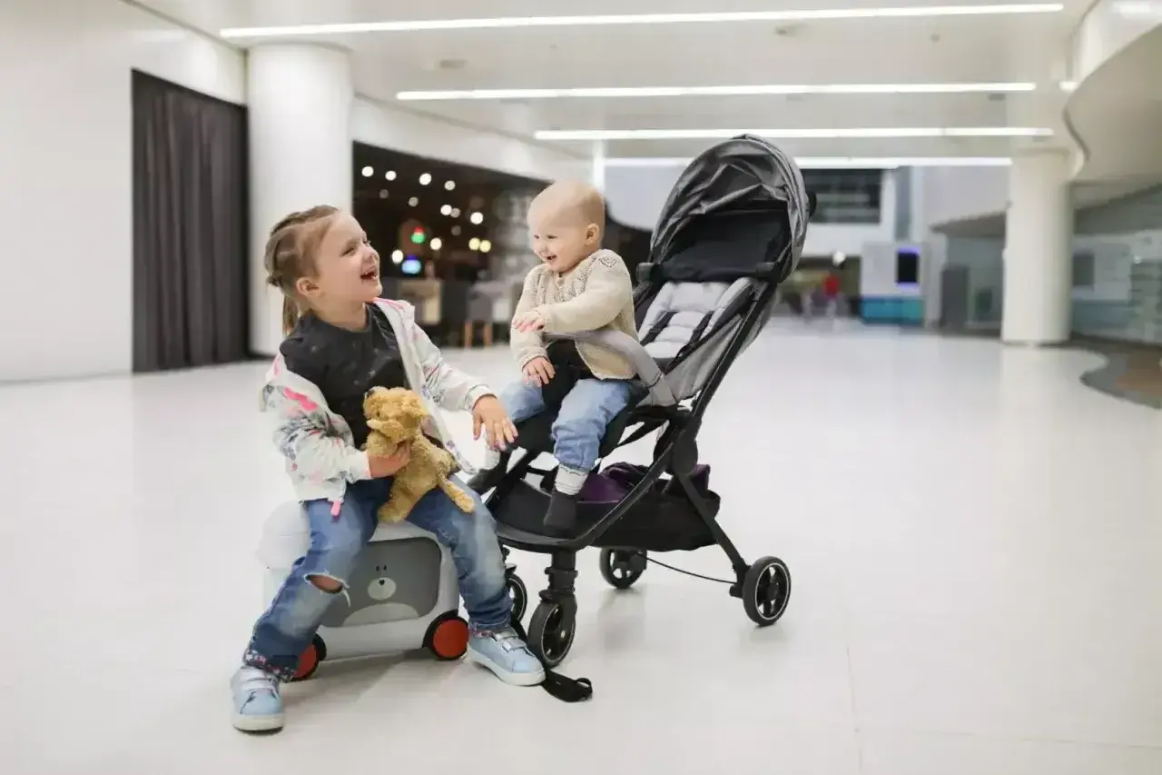 Dwie dziewczynki, jedna siedzi na walizce, druga w wózku, gotowe na podróż z Ryanair. Wózek dla 3 latka idealny na lotnisko.