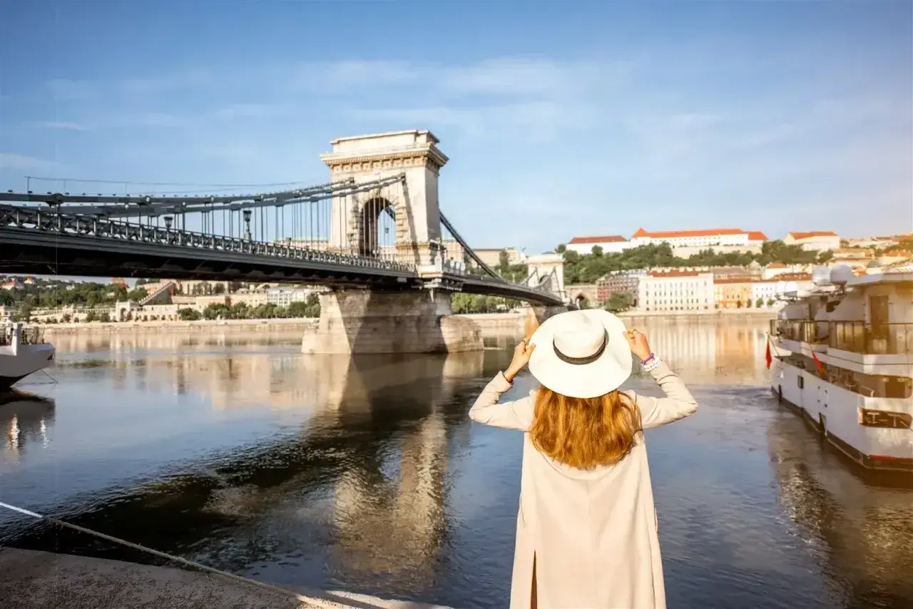 Kobieta w kapeluszu podziwia Most Łańcuchowy w Budapeszcie. W tle widok na Dunaj i budynki, a w portfelu czeka budapeszt waluta na dalsze zwiedzanie.