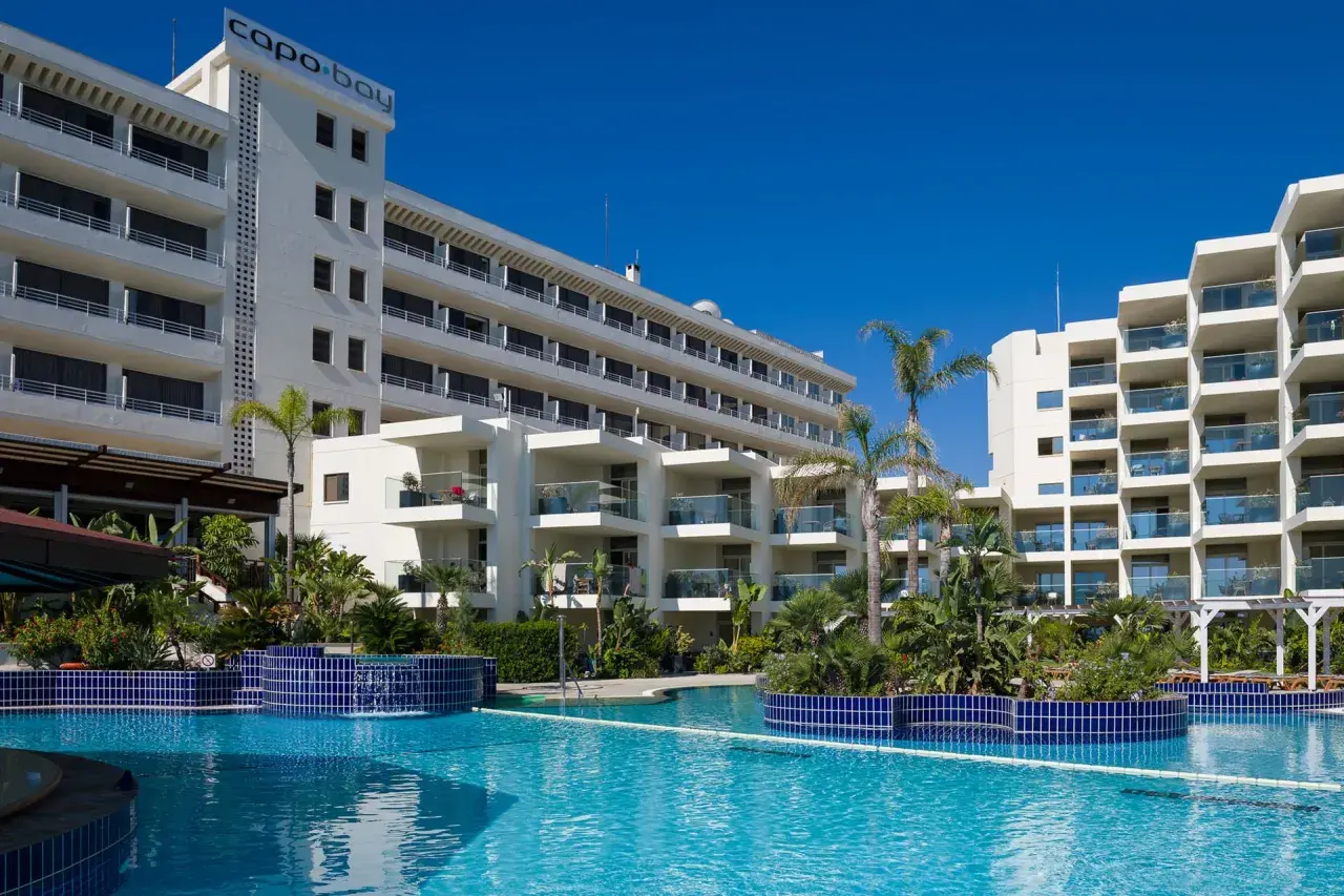Basen hotelowy Capo Bay w Protaras z palmami i białymi budynkami hotelowymi pod błękitnym niebem.