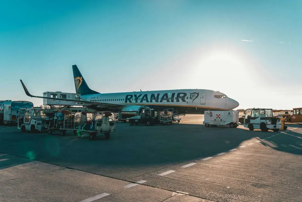 Samolot Ryanair na płycie lotniska, przygotowywany do lotu. Widać wózki z bagażem i sprzęt obsługi naziemnej.