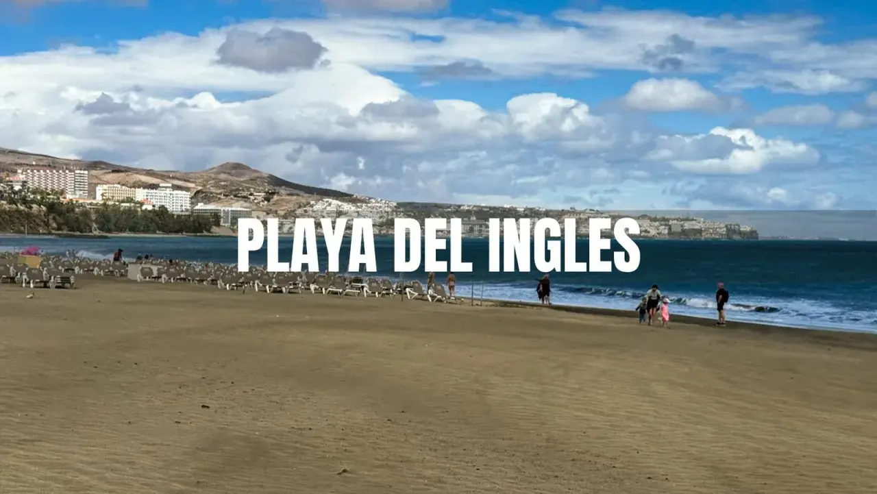Piękna plaża Playa del Inglés z rzędami leżaków i kilkoma spacerującymi ludźmi. W tle widać hotele i góry.