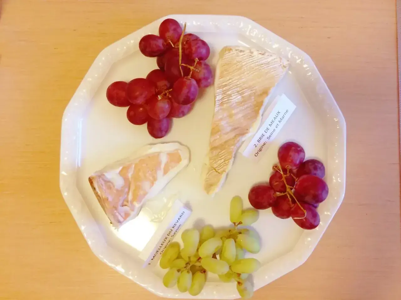 Talerz z serami Brie de Meaux i Moelleux du Revard, winogronami i etykietami, idealny do degustacji kuchni francuskiej.