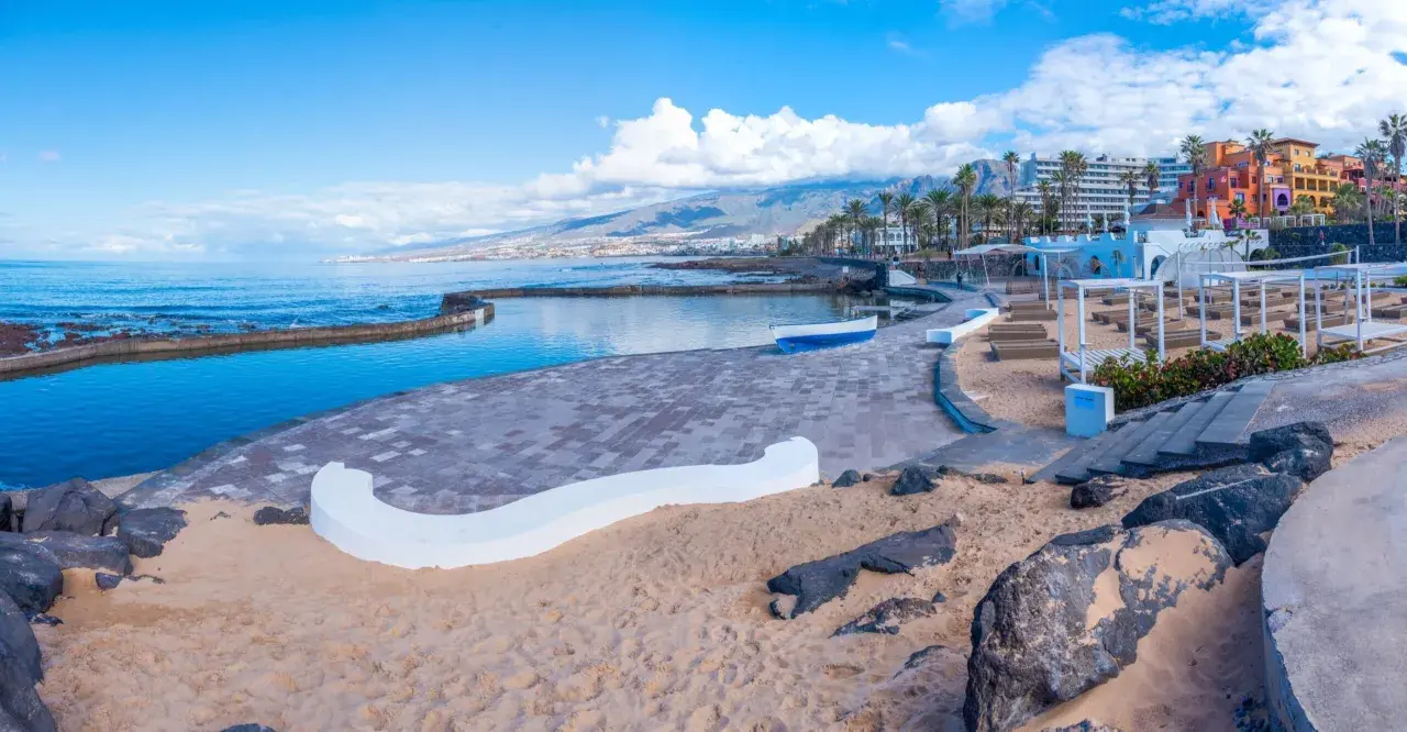 Słoneczna playa de las americas z basenem, piaskiem i kolorowymi budynkami w tle.