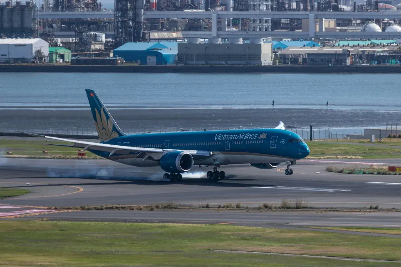 Błękitny 787 Dreamliner Vietnam Airlines ląduje, unosząc dym z opon na pasie startowym. W tle widać przemysłowy krajobraz i wodę.