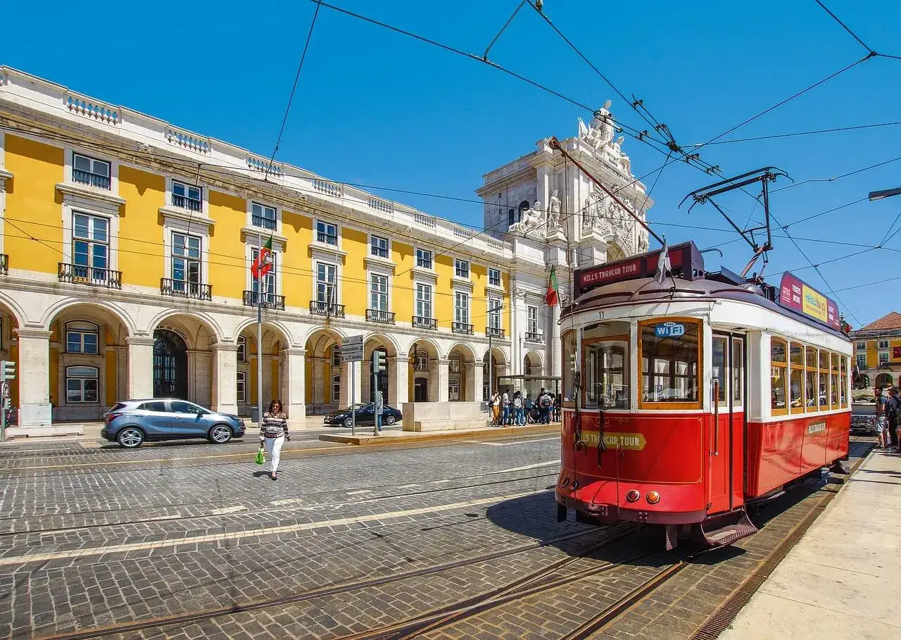 Czerwony tramwaj w Lizbonie, obok żółtego budynku i łuku triumfalnego. Czy do Portugalii potrzebny jest paszport? Zależy od Twojego obywatelstwa.