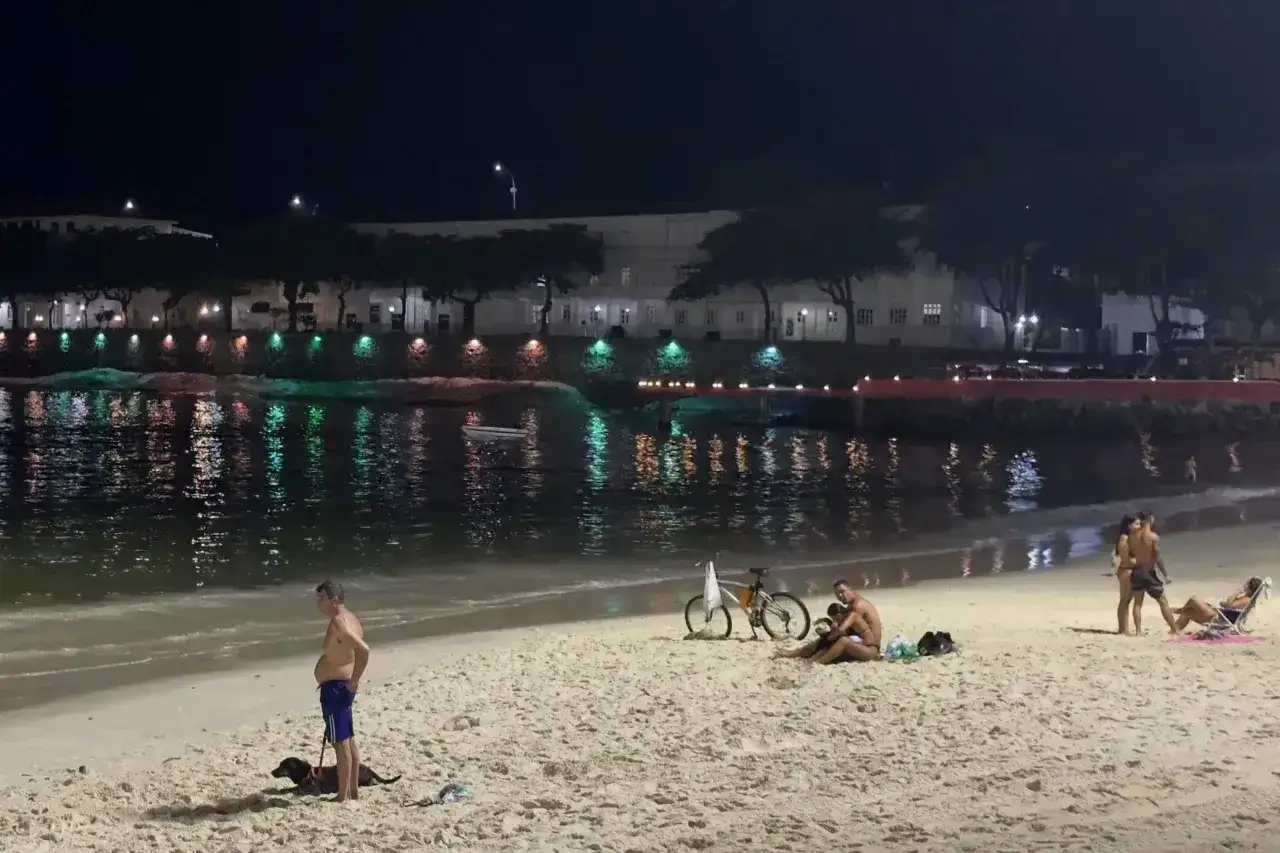 Nocna plaża w Brazylii, ludzie odpoczywają, psy spacerują. Czy Brazylia jest bezpieczna? Widać spokojną atmosferę, światła miasta odbijają się w wodzie.