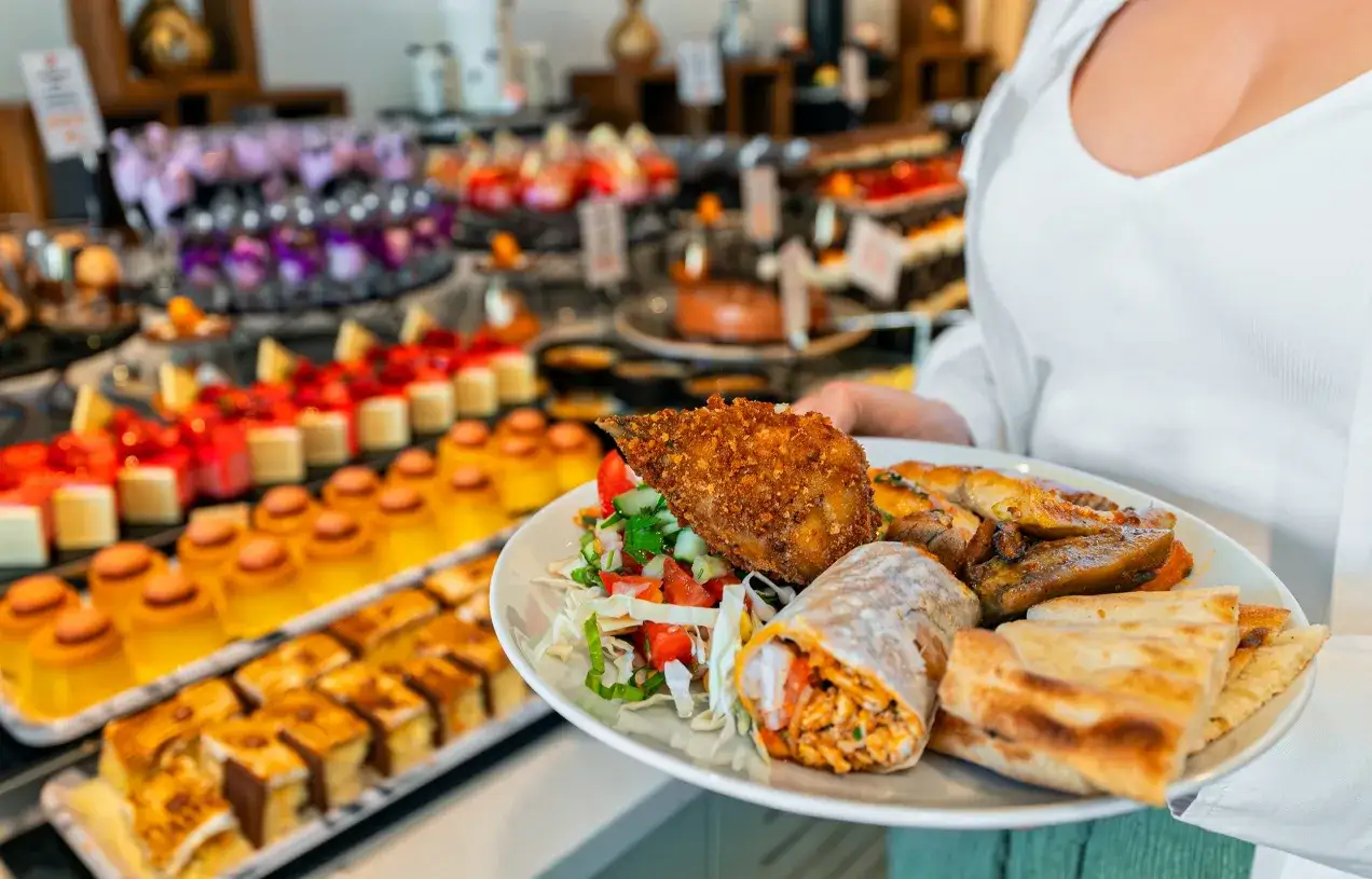 Talerz pełen smakołyków: panierowany kurczak, wrap, warzywa i pieczywo. W tle kuszący bufet z deserami i daniami, prawdziwy full board plus.