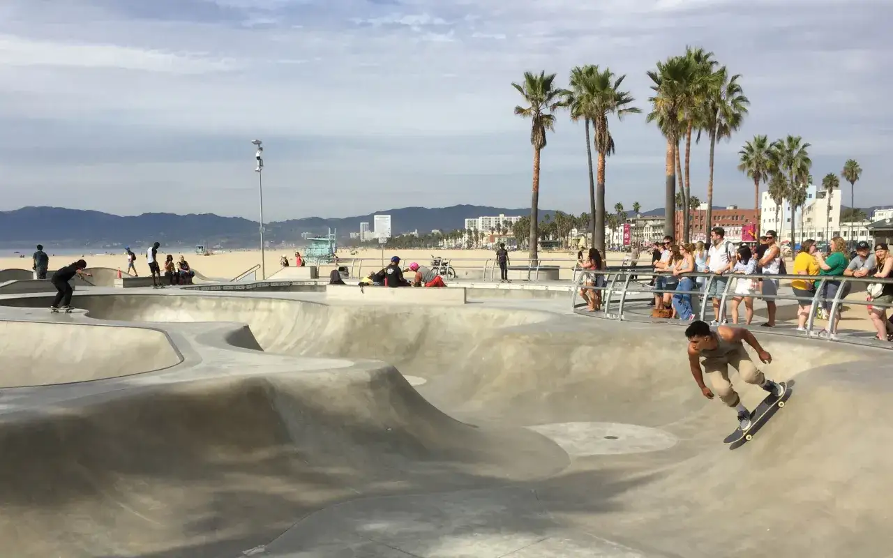 Na Venice Beach młody mężczyzna na deskorolce zjeżdża po betonowej rampie, podczas gdy tłum obserwuje go zza barierek.