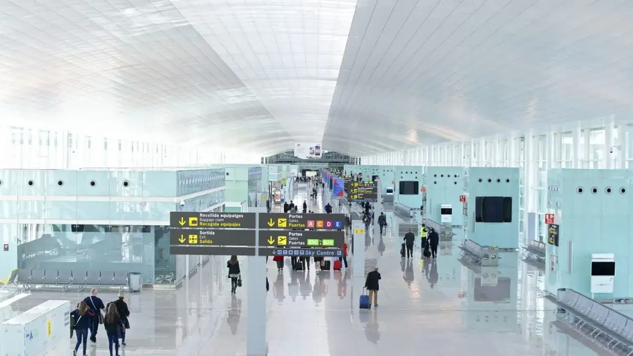 Nowe, jasne wnętrze terminalu lotniska Barcelona El Prat. Pasażerowie z bagażami przemieszczają się pod nowoczesnym sufitem i wśród szklanych konstrukcji.