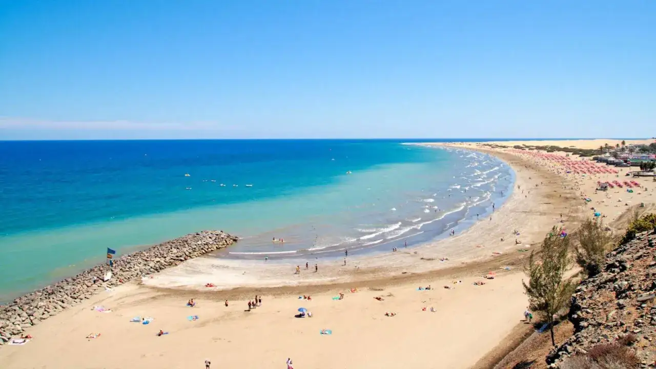 Słoneczna plaża Playa del Inglés z turkusową wodą, kamiennym falochronem i licznymi leżakami.