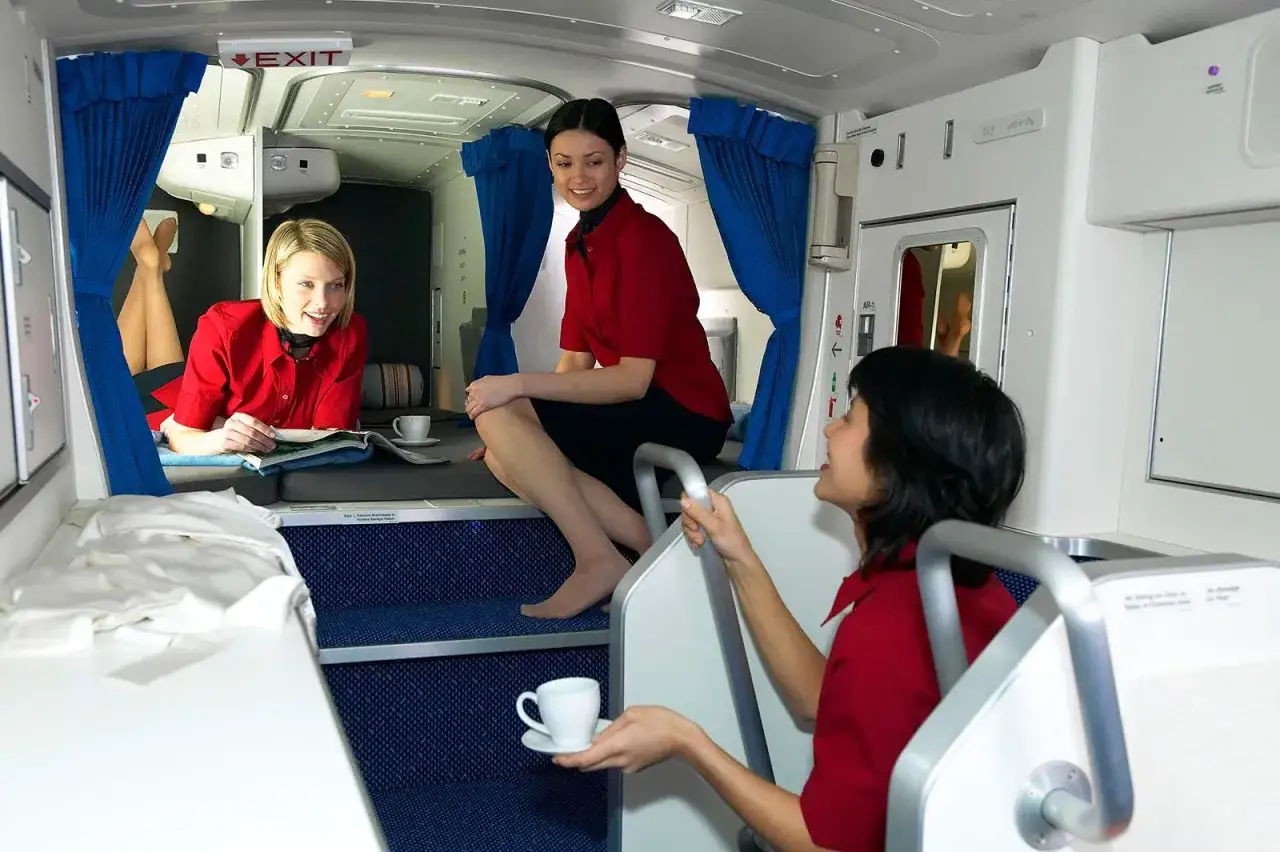 Trzy stewardessy w czerwonych uniformach odpoczywają w kabinie samolotu. Jedna leży, czytając, druga siedzi, a trzecia podaje kawę. To ich praca.
